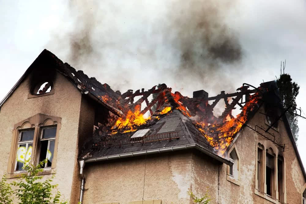 A large house with a partially collapsed roof is engulfed in flames, thick smoke billowing into the sky. While the stone walls remain intact, water damage restoration will be crucial once the fire-consuming wooden roof structure is extinguished. Sparse greenery surrounds the building.