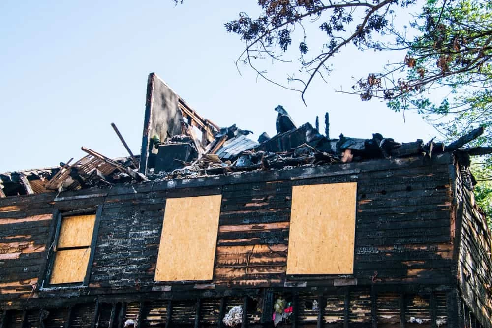 A burned house with its roof partially collapsed shows the effects of fire damage. The charred exterior walls and boarded-up windows await restoration. Nearby branches extend into view against a clear blue sky, emphasizing the urgent need for expert fire damage restoration.