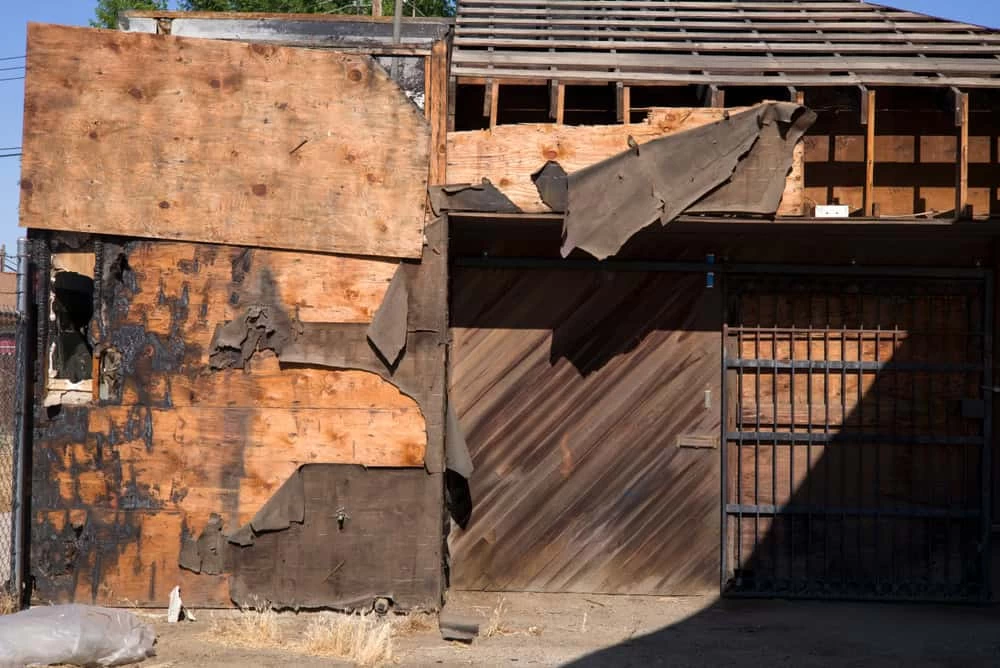 A weathered building with wooden panels and peeling tar paper on the exterior, shows signs of fire damage. Some sections are charred, hinting at the need for restoration. A metal gate stands before a wooden door, as sunlight casts shadows on the concrete ground.