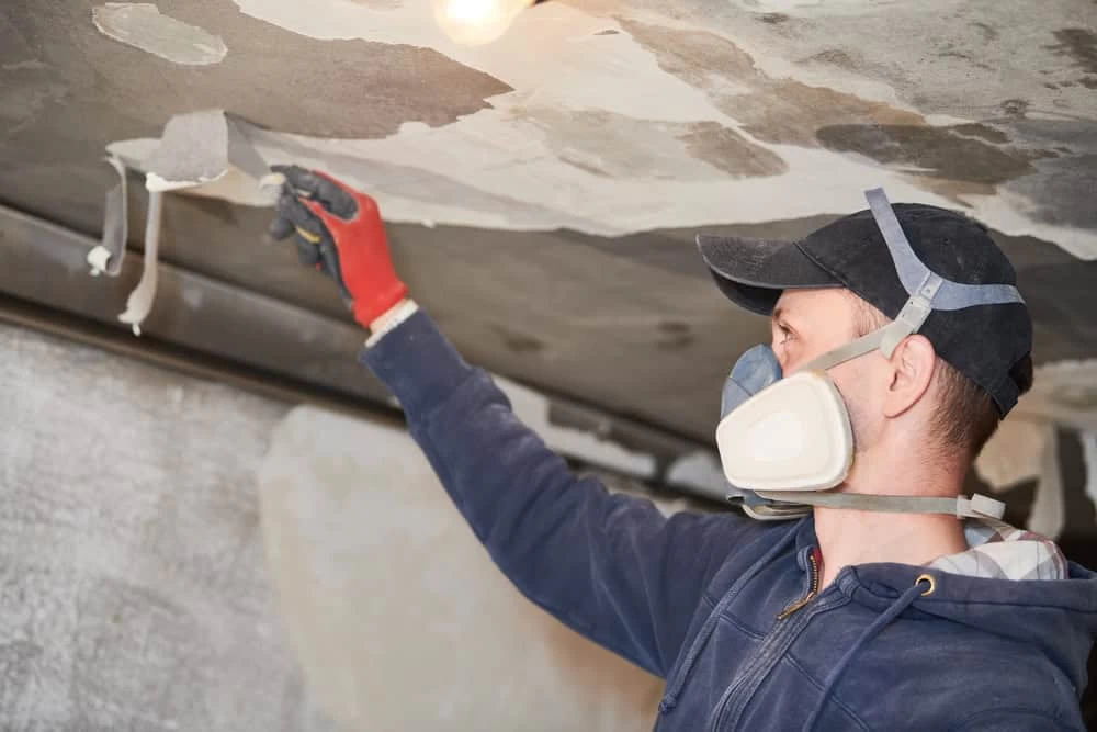 A person wearing a cap, mask, and gloves is removing old paint or plaster from a ceiling using a scraping tool. This meticulous work is part of a restoration company's efforts to revive the space, with a light bulb casting some clarity on the transformation underway.