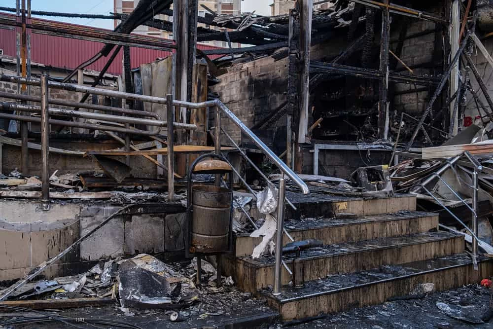 Image of a building extensively damaged by fire, showing charred debris, twisted metal, and blackened walls. Steps lead to a partially destroyed entrance amidst rubble. A damaged railing and trash can are also present. Perfect for showcasing the expertise of a restoration company in rebuilding from ruins.