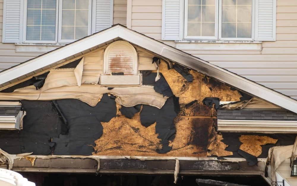The image shows the front of a house with fire damage. The upper section has charred and peeling siding, exposing the underlying material. Windows with white shutters are intact above the damaged area, highlighting the need for expert Fire Damage Restoration to restore its former glory.