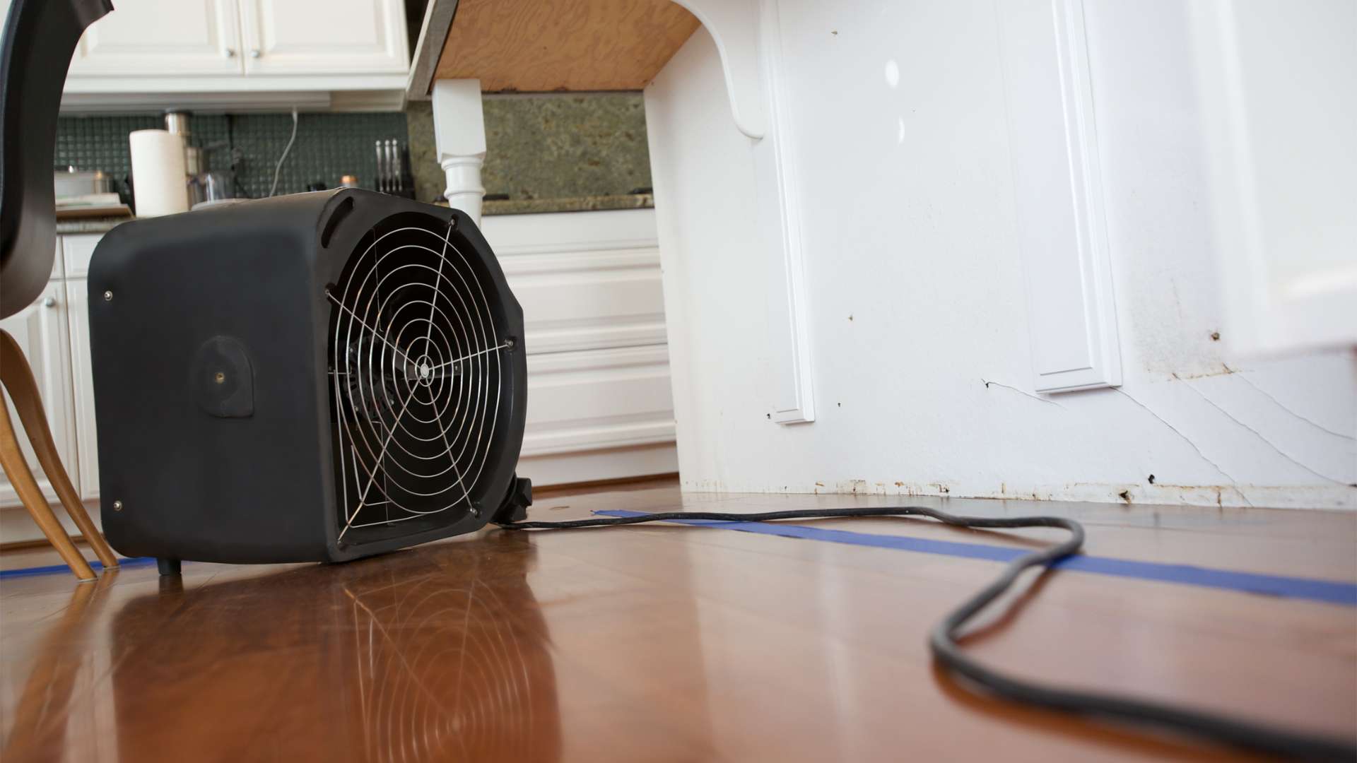 A black industrial fan sits on a wooden floor, working tirelessly for water damage restoration services in the corner beneath a kitchen counter. Its power cord snakes across the floor, with white cabinets and part of a chair visible in the background.