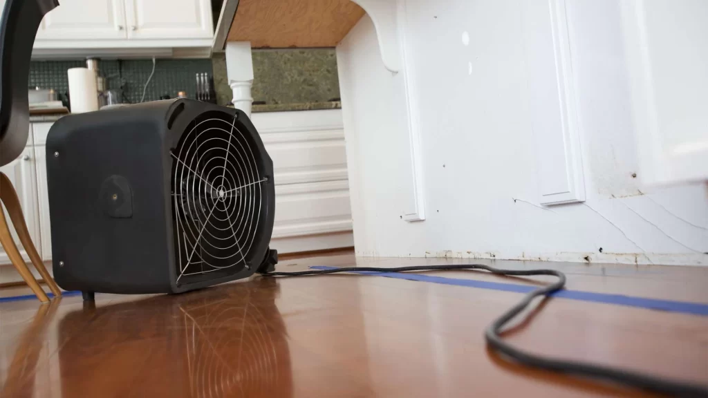 A black industrial fan sits on a wooden floor, working tirelessly for water damage restoration services in the corner beneath a kitchen counter. Its power cord snakes across the floor, with white cabinets and part of a chair visible in the background.