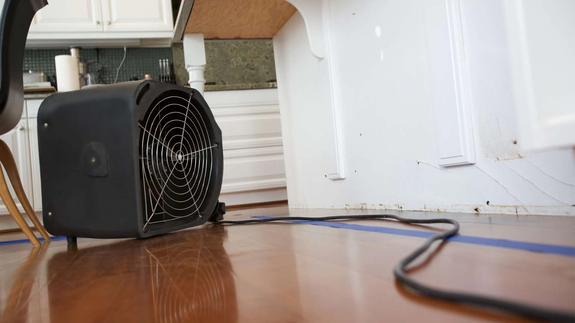 A black industrial fan sits on a wooden floor, working tirelessly for water damage restoration services in the corner beneath a kitchen counter. Its power cord snakes across the floor, with white cabinets and part of a chair visible in the background.