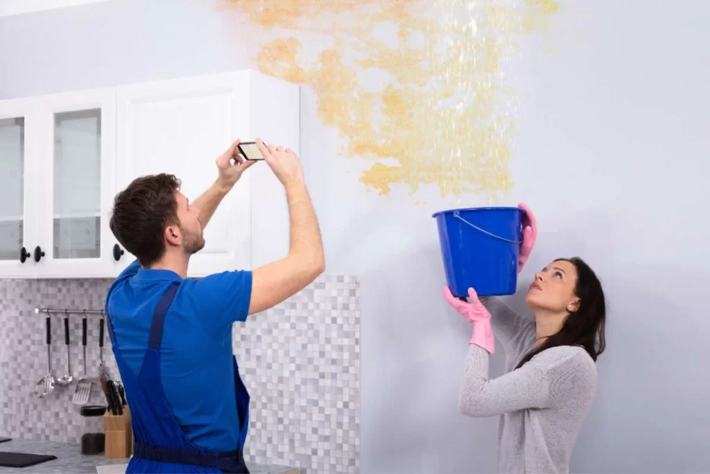 A woman in pink gloves holds a blue bucket under a kitchen ceiling leak, signaling the need for water damage restoration. A man in a blue shirt and overalls documents the yellow-stained ceiling with his phone, ready to call in professional water damage restoration services. Kitchen cabinets can be glimpsed nearby.