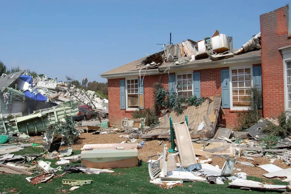 A red brick house with severe roof damage and debris scattered around serves as a somber testament to recent chaos. The lawn, littered with wreckage, hints at a need for natural disaster restoration. A clear sky offers a stark contrast to this scene of destruction.