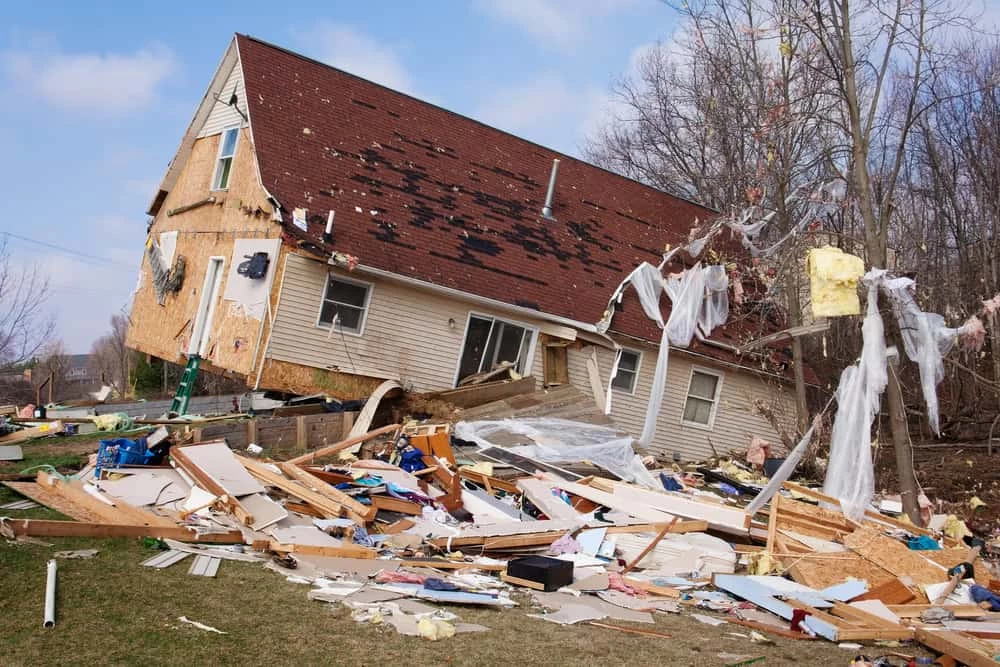 A house leans to one side with extensive structural damage, surrounded by debris including wood, insulation, and household items scattered across the lawn. Nearby trees are bare, and the sky is partly cloudy, highlighting the urgent need for natural disaster restoration.