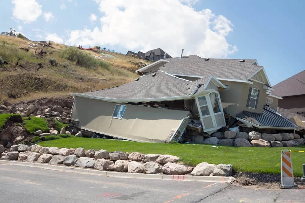 A house is partially collapsed and leaning on rocks at the edge of a sloped hill, showing signs of severe structural distress likely caused by water damage. In the foreground, there's a small road with construction markers. Clouds loom in the sky, hinting at more challenges ahead for restoration efforts.
