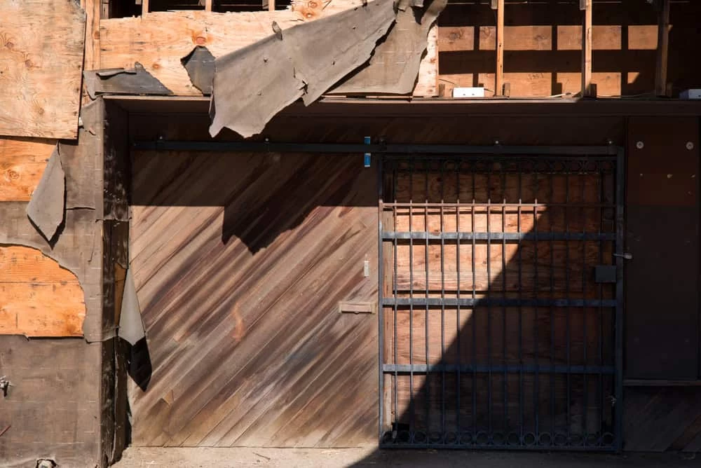 A weathered, boarded-up building facade with partially torn plywood and exposed wooden planks suggests the need for natural disaster restoration. A rusty metal gate is in the center, casting a shadow on the wall. Debris and loose materials are visible above the gate.