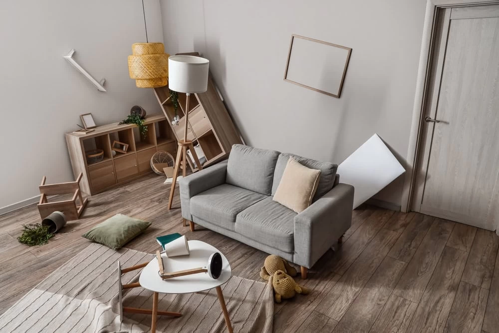 A room in disarray, with overturned furniture including a gray sofa and fallen side tables. A tilted floor lamp and toppled cabinet hint at chaos, while a rolled carpet on the wooden floor suggests recent water damage requiring restoration.