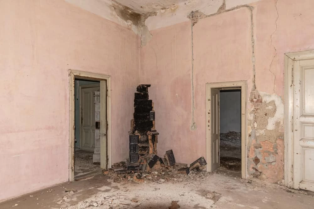 An abandoned room with peeling pink walls and a deteriorating ceiling hints at past neglect. In the center, a crumbling, blackened stove structure lies in disarray. Doorways reveal more neglected spaces, ripe for the expertise of a restoration company skilled in mold removal and water damage restoration services.