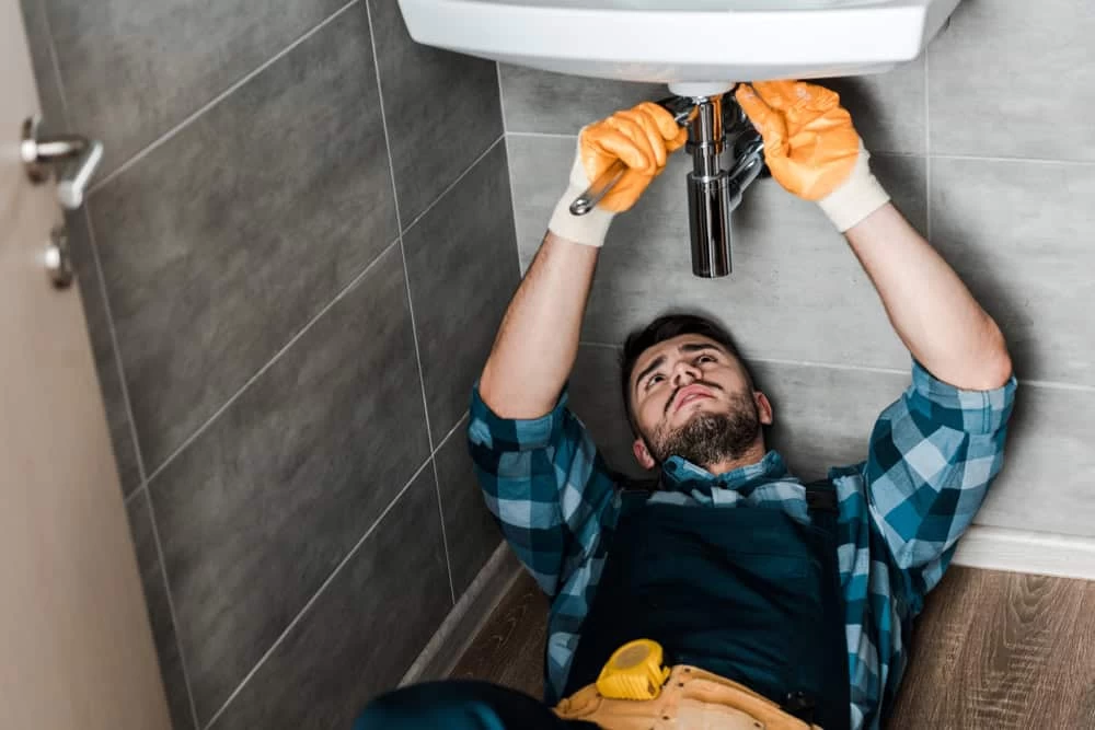 A plumber in a checkered shirt and overalls lies on the floor, wearing orange gloves, expertly working on piping under a sink in a tiled bathroom. His skills are crucial for preventing future issues, just like water damage restoration services mitigate further problems after leaks.