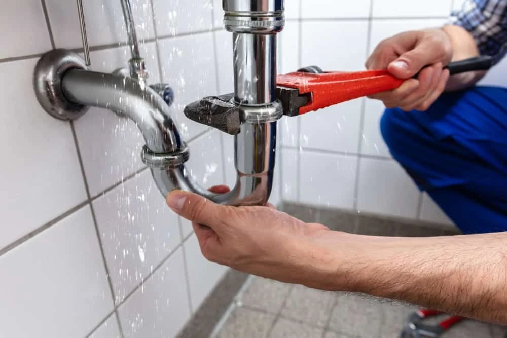 A person is using a red pipe wrench to fix a leaky pipe under a sink, preventing potential water damage. Water sprays from the connection, surrounded by white tiled walls and a gray tiled floor.