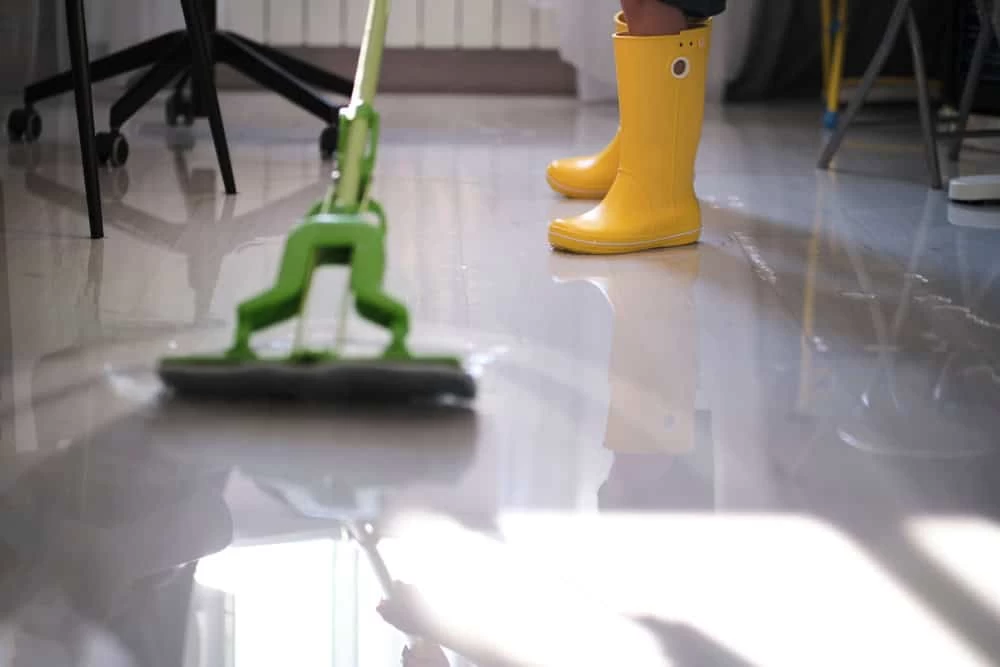A person in yellow rain boots mops a shiny floor, reflecting the expertise of a Restoration Company. The green mop moves skillfully amidst the office furniture in this bright, indoor scene.