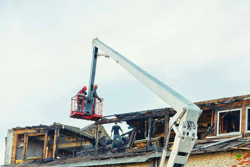 Two construction workers in a lift basket inspect and repair a partially damaged building. The structure shows signs of fire damage, with burnt wood and debris. As part of a restoration company, they focus on natural disaster restoration. The sky is overcast, and the workers wear safety helmets and harnesses.