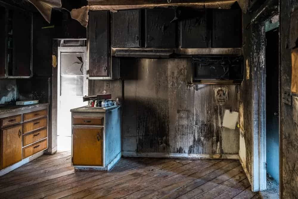 A kitchen in CA with burnt, blackened walls and ceiling, damaged cabinets, and a dirty wooden floor shows significant fire and smoke damage. Light shines through a doorway—ideal for restoration services Ventura County can trust.