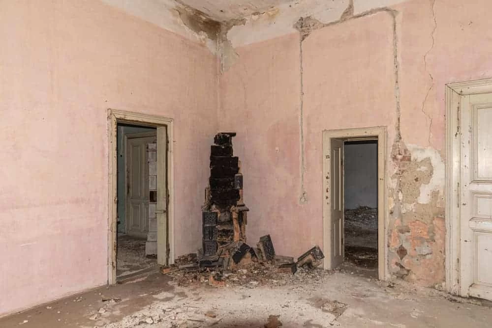 An abandoned room in CA with peeling pink walls, crumbling plaster, and a broken, charred stove in the corner. Two doorways and a closed white door are visible amid debris—perfect for restoration services Ventura County can offer.