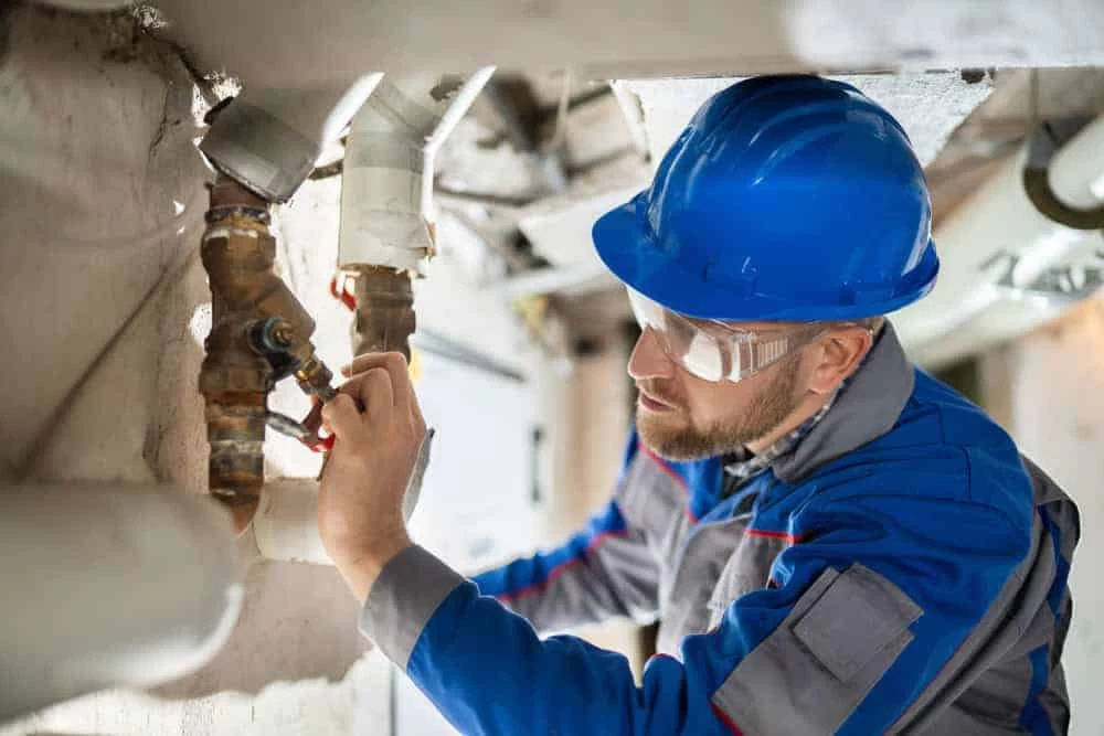 A plumber wearing a blue hard hat, safety glasses, and a work uniform adjusts a valve on a pipe in a basement, demonstrating the expertise found in restoration services Ventura County, CA.